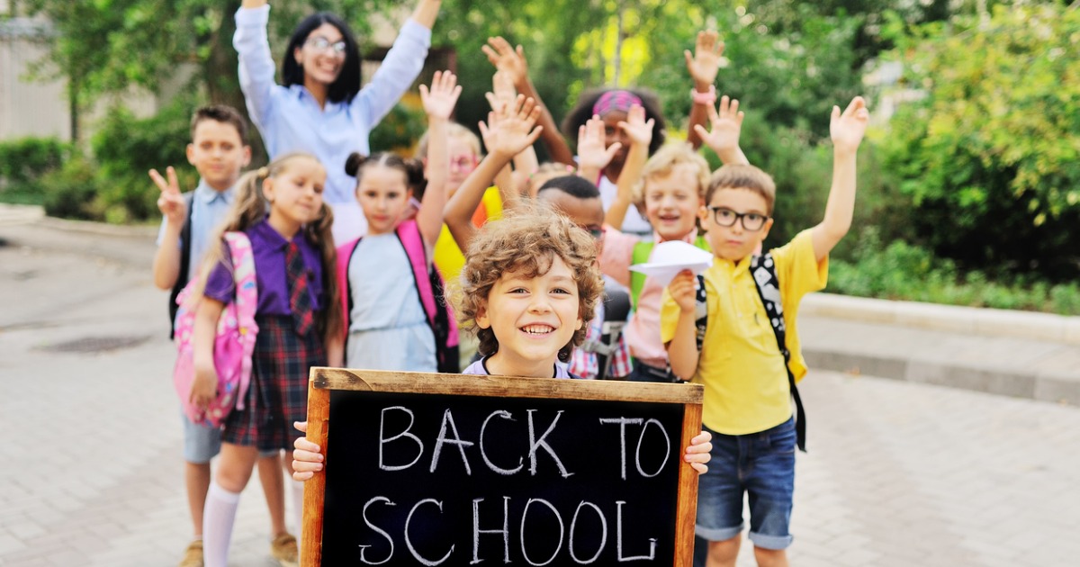 School age boy holds "Back to School" sign while friends and parents wave in the background