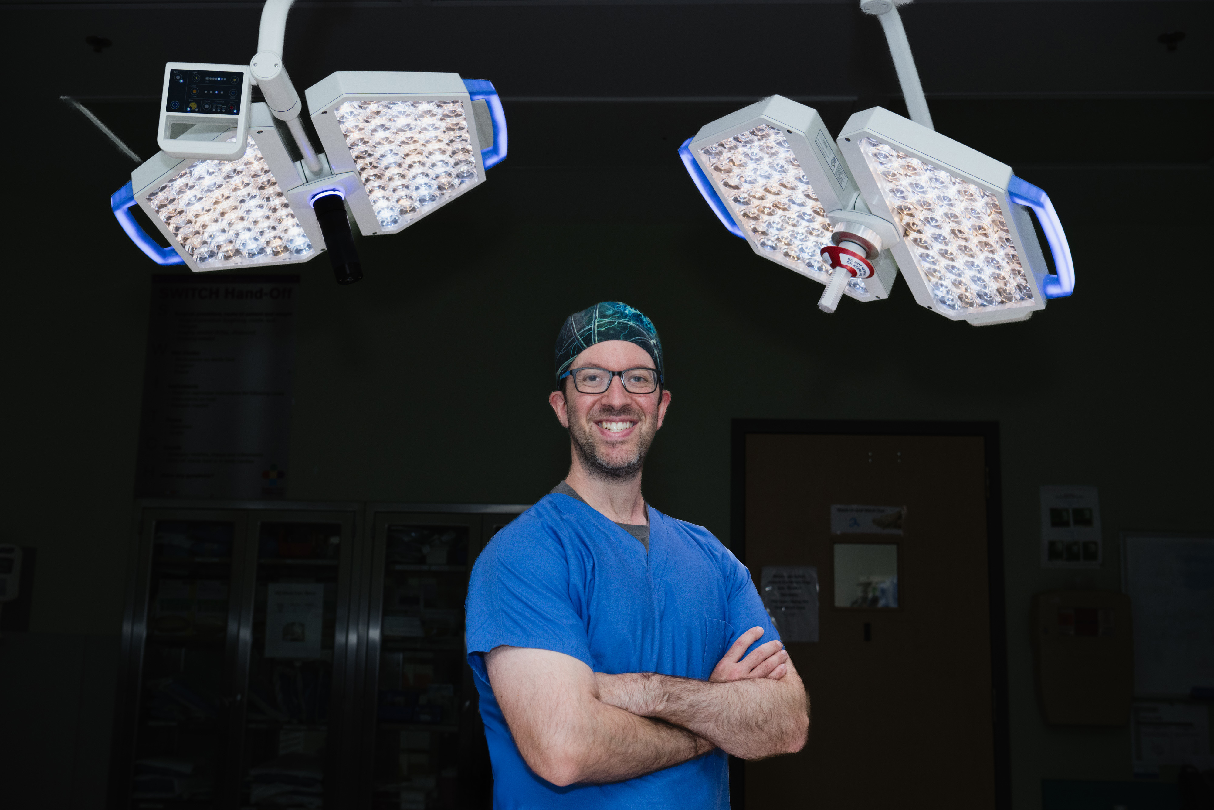 Dr. Hersh, neurosurgeon, stands in the operation room with is arms crossed, with operating room lights behind him.