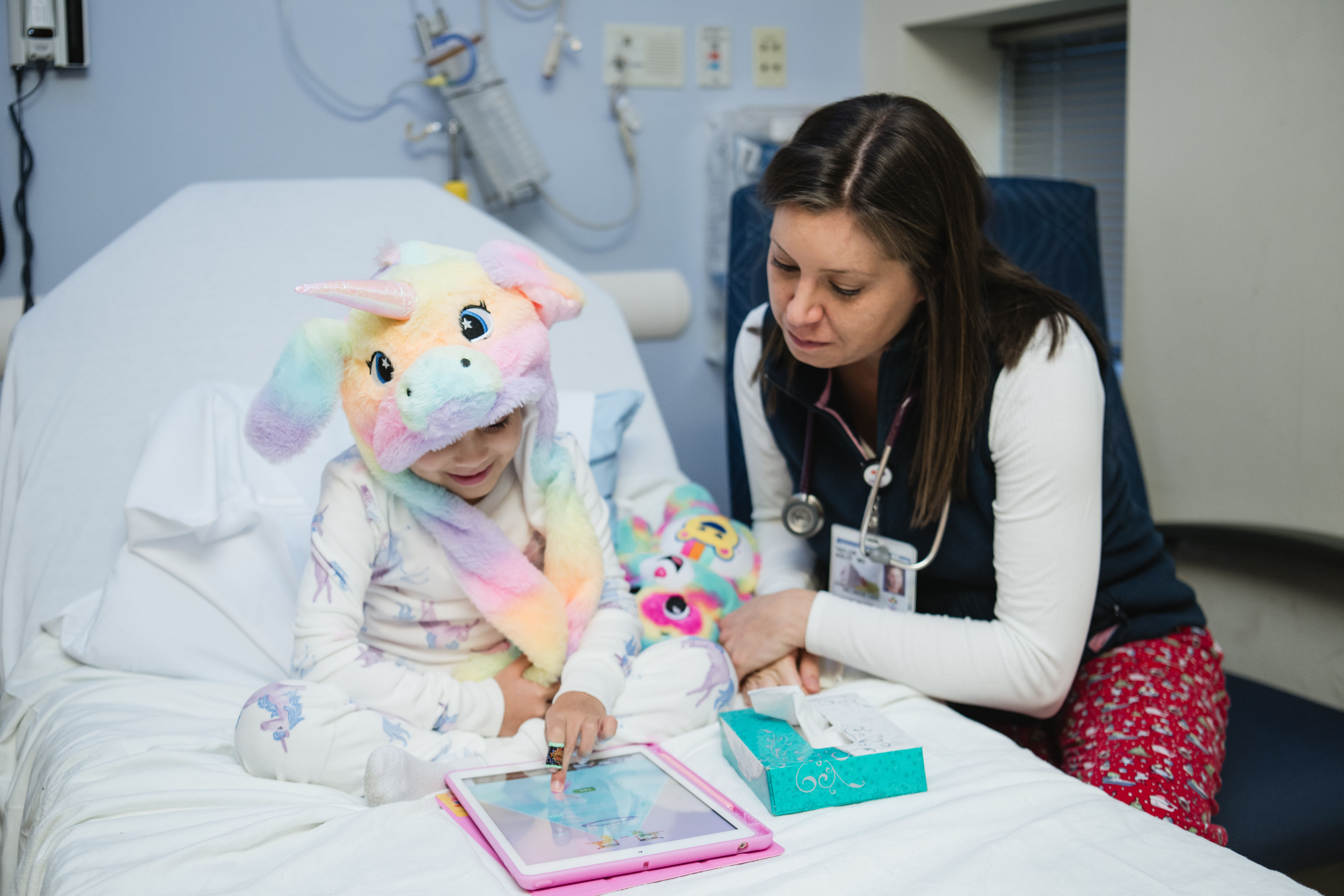 A hem-onc patient seated in a hospital bed shows a doctor her i-pad. A hem-onc patient seated in a hospital bed shows a doctor her i-pad.