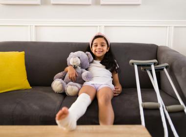 School-aged girl on couch holding stuffy, crutches at side. Wearing leg cast.