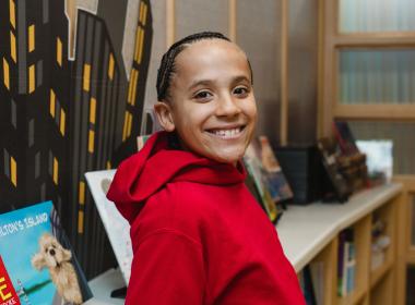 School-aged boy smiling in library, wearing red sweatshirt