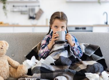 Sick girl with flu drinking from mug sitting on couch next to teddy bear