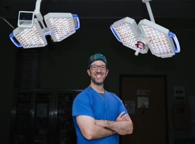 Dr. Hersh, neurosurgeon, stands in the operation room with is arms crossed, with operating room lights behind him. Dr. Hersh, neurosurgeon, stands in the operation room with is arms crossed, with operating room lights behind him.