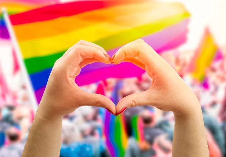 Supporting hands make heart sign and wave in front of a rainbow flag flying on the sidelines of a summer Pride parade