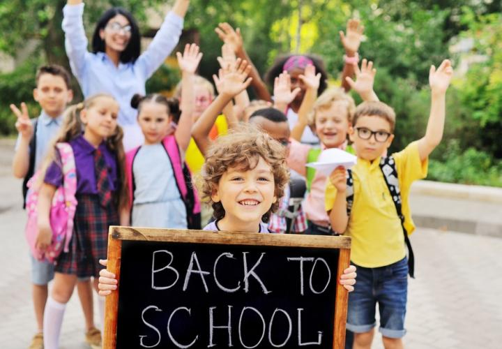 School age boy holds "Back to School" sign while friends and parents wave in the background