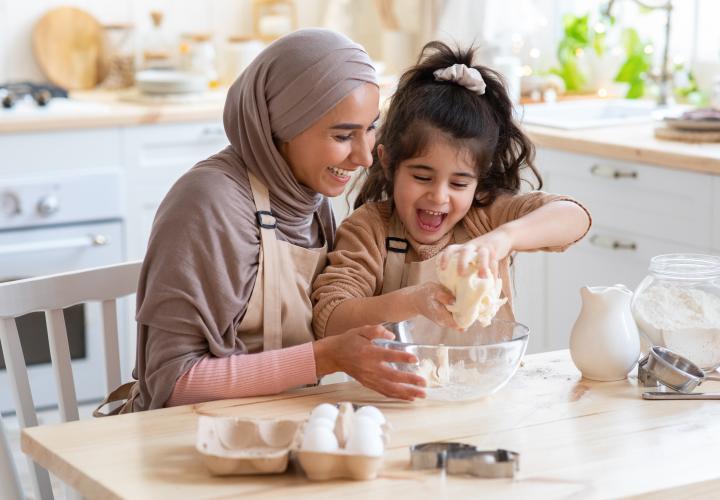 Mom and young daughter crack eggs into a bowl in kitchen, happily.