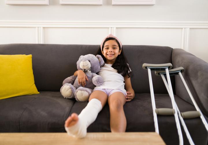 School-aged girl on couch holding stuffy, crutches at side. Wearing leg cast.