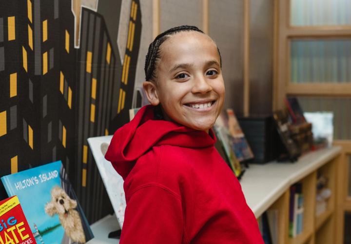 School-aged boy smiling in library, wearing red sweatshirt