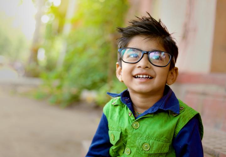Young boy, smiling, looking up. Wearing glasses and green and blue jacket.