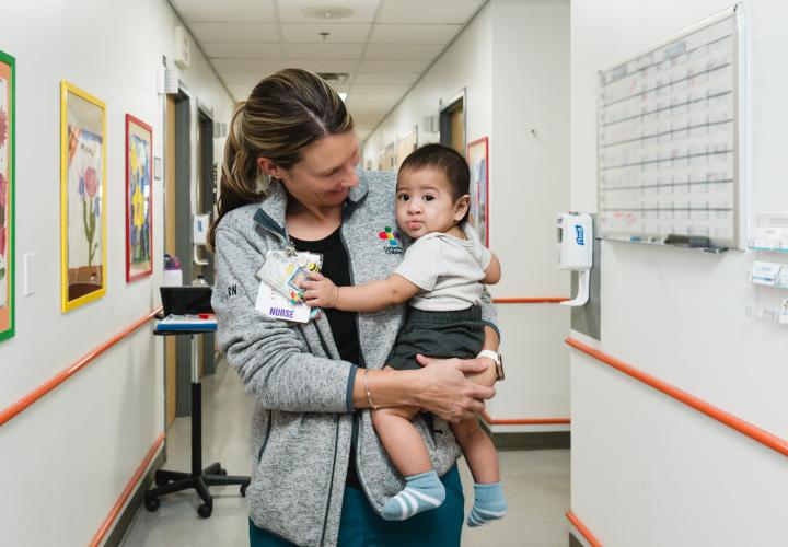 A nurse holds an infant craniofacial patient in the clinic hallway.
