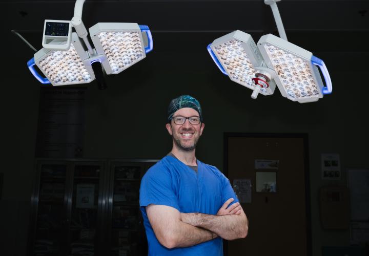 Dr. Hersh, neurosurgeon, stands in the operation room with is arms crossed, with operating room lights behind him.