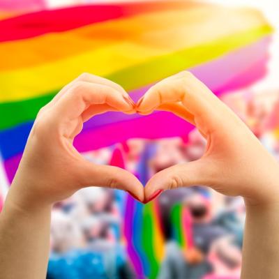 Supporting hands make heart sign and wave in front of a rainbow flag flying on the sidelines of a summer Pride parade