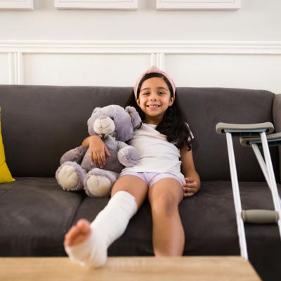 School-aged girl on couch holding stuffy, crutches at side. Wearing leg cast.