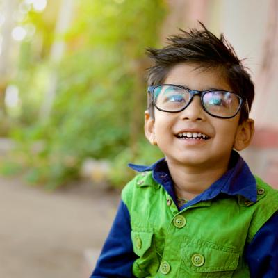 Young boy, smiling, looking up. Wearing glasses and green and blue jacket.