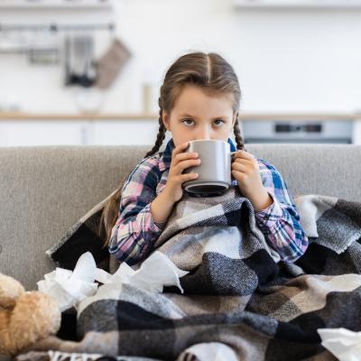 Sick girl with flu drinking from mug sitting on couch next to teddy bear